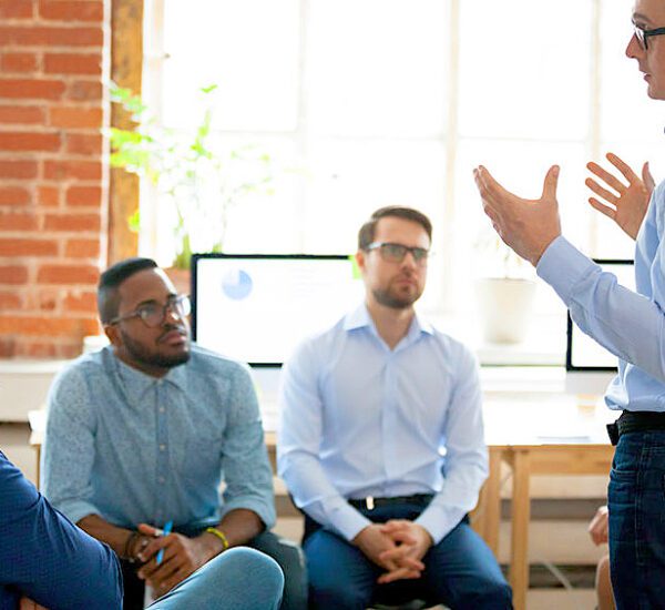 Confident male leader mentor coach speaking at diverse team meeting, presenter speaker manager explaining new business plan strategy to employees group during office briefing at corporate training